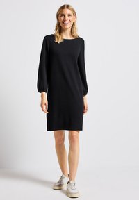 Robe noire à manches longues avec un décolleté carré et des poignets élastiques, en tissu tricot doux, portée avec des baskets légères. Silhouette simple et droite.