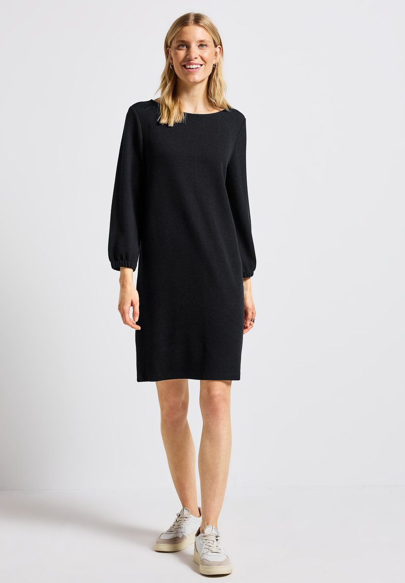 Robe noire à manches longues avec un décolleté carré et des poignets élastiques, en tissu tricot doux, portée avec des baskets légères. Silhouette simple et droite.