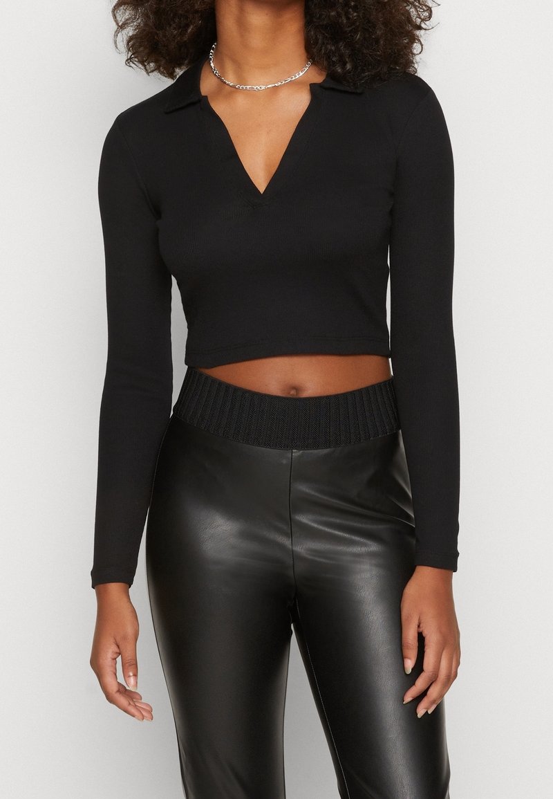 Schwarzes langärmliges Cropped-Top mit Kragen und V-Ausschnitt, kombiniert mit hochgeschnittenen schwarzen Kunstlederhosen mit strukturiertem Taillenbund.