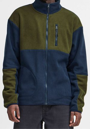 Mann trägt eine halb olivgrüne, halb marineblaue Fleecejacke mit Reißverschlusstaschen vorne und auf der Brust, steht mit entspannten Händen an den Seiten.