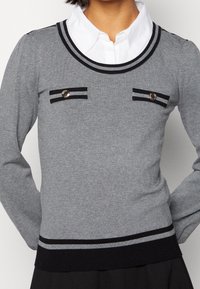 Pull en tricot gris avec un col arrondi, des accents à rayures noires et des boutons métalliques sur les poches poitrines. Design ajusté avec des manches longues.