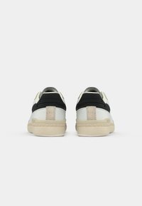 Des baskets blanches et noires avec un accent en daim, une forme arrondie et une semelle en caoutchouc texturée, dotées d'une étiquette arrière et d'éléments de design minimalistes.