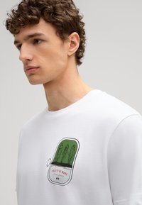 Weißes Baumwoll-T-Shirt mit einem bunten Grafikdesign eines Fischlabels, das grüne und rote Akzente sowie eine glatte Textur aufweist.