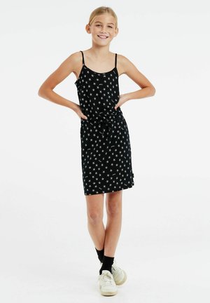 Zwart jurkje met wit sterrenpatroon, voorzien van dunne bandjes, een strik taille en een rok tot op de knie. Draag het met witte sneakers en zwarte sokken.