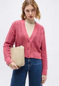 Νεαρή γυναίκα με ροζ πλεκτή ζακέτα πάνω από μια κρεμ ριμπ μπλούζα, κρατώντας μια μπεζ δερμάτινη τσάντα, ντυμένη με μπλε τζιν.