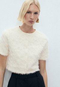 Kortärmad, ljus cremefärgad kofta gjord av texturerat, fluffigt material med rund halsringning och framknäppning.