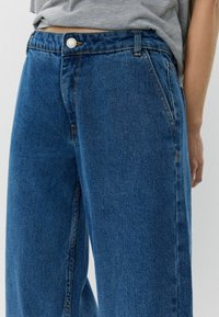Pantalones vaqueros de denim azul de pierna ancha con una textura suave, que cuentan con un cierre de botón en la parte delantera, presillas para cinturón y bolsillos laterales.