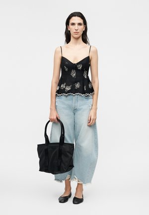 Vrouw draagt een zwarte spaghetti-band top met bloemenprint, lichte wijde gescheurde jeans, zwarte platte schoenen en houdt een zwarte tote tas vast.