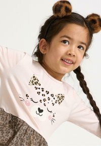 Maglietta lunga a maniche rosa chiaro con un design di faccia di gatto stampato a leopardo, abbinata a una gonna di tulle a stampa leopardata marrone. Cerchietto con motivo animale marrone.