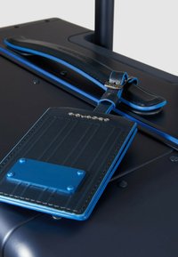 Etichetta per bagaglio blu scuro attaccata alla maniglia di una valigia. L'etichetta presenta una superficie testurizzata, un accento blu a contrasto e una fibbia metallica ad anello.