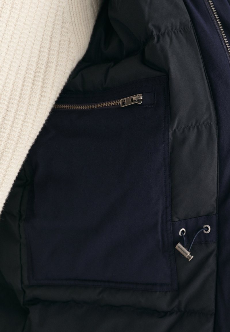 Interno di giacca della marina con tasca con zip e cordoncino regolabile, indossata sopra un maglione lavorato a maglia color crema.