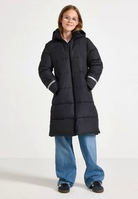 Schwarzer, gepolsterter langer Mantel mit Kapuze, ausgestattet mit einem Reißverschluss vorne und grauen Akzenten an den Bündchen. Getragen über weitem, blauen Jeans und schwarzen Sneakers.