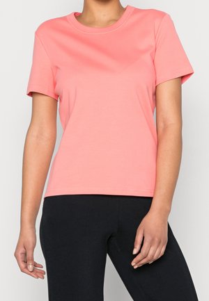 Mujer vistiendo una camiseta coral lisa de manga corta y cuello redondo, y pantalones ajustados negros, de pie contra un fondo claro.