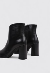 Botas de tobillo de cuero negro con una textura suave, punta redondeada y tacones cilíndricos. Cierre lateral con cremallera para ponerlas y quitarlas fácilmente.