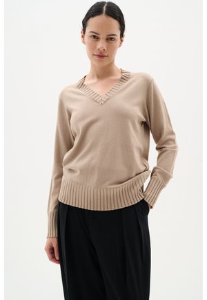 Maglione lavorato a maglia beige con scollatura a V, polsini e orlo a costine, dalla texture liscia e dalla vestibilità morbida, abbinato a pantaloni scuri.
