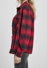 Kvinde iført en rød og sort ternet skjorte med knapper og mørkegrå jeans, stående sidelæns imod en ensfarvet lys grå baggrund.