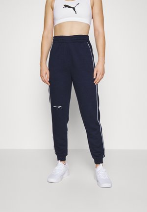 Osoba nosząca granatowe spodnie dresowe Reebok z białymi paskami i białe buty sportowe, połączone z białym sportowym biustonoszem Puma.
