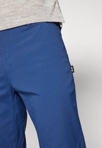 Blue trousers made of smooth fabric, featuring a front pocket and a small black label on the side. The design is slim-fit with minimal detailing.