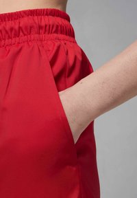 Rote Sportshorts mit einem elastischen Bund, ausgestattet mit einer Seitentasche und glattem, strukturiertem Stoff. Fokussiert auf Passform und Praktikabilität.
