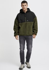 Fleecemuddad svart och olivgrön huppartröja med halvdragkedja, texturerat tyg och en liten logotyp på framsidan.