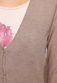 Cárdigan de punto color marrón claro con escote en V y cierre de botones, superpuesto a una camisa rosa pálido con un diseño gráfico floral.