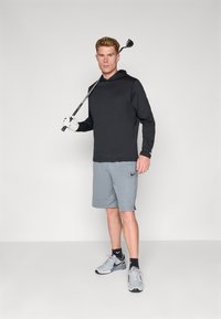 Svart langermet hettegenser og grå shorts. Golft klubb holdt med hanskekledd hånd. Grå joggesko med svarte detaljer, stående på en ensfarget bakgrunn.