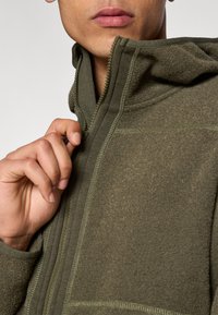 Ζακέτα φλις με φερμουάρ σε σκούρο πράσινο, με υφή υφάσματος, ορατές λεπτομέρειες ραφής και ψηλό γιακά. Το χέρι του μοντέλου ρυθμίζει το φερμουάρ.