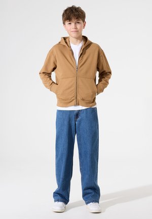 Haut à zip marron avec une coupe décontractée, grandes poches avant, associé à un jean bleu ample et des baskets blanches. Texture de tissu lisse.