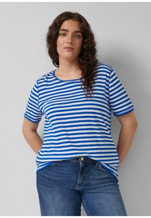 Frau mit lockigem dunklem Haar, die ein blau-weiß gestreiftes Kurzarmhemd und blaue Jeans trägt, steht vor einem hellgrauen Hintergrund.