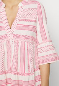 Blouse imprimée en tissu léger, avec des motifs géométriques roses et des rayures, encolure en V, et manches courtes à volants avec détails froncés.