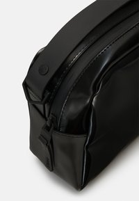 Černá, lesklá kosmetická taška s hladkým povrchem, obdélníkovým tvarem a pevným uchem. Obsahuje černé kovové prvky a detail loga.
