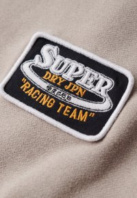Beige stof med et sort og hvidt broderet mærke. Mærket har teksten "SUPER DRY JPN" og "RACING TEAM" i orange.