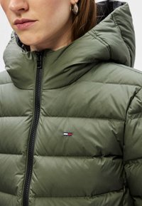 Chaqueta acolchada verde con un cuello alto, cremallera negra y bolsillos laterales; cuenta con un pequeño logotipo bordado con acentos en rojo, blanco y azul.