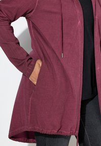 Burgunderfarvet lang cardigan med hætte, struktureret stof, åben front, sidelommer og en samlet kant. Sammen med en sort top og mørke jeans.