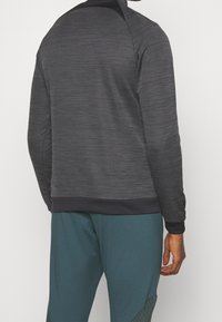 Černá sportovní mikina s texturovaným vzorem, žebrovanými manžetami a lemem a vysokým límcem. V kombinaci s tyrkysovými kalhotami s šikmým designem.