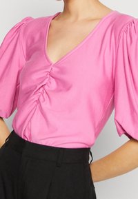 Růžový krátký rukáv top s hlubokým výstřihem do V a řaseným detailem na přední straně. Hladká textura a mírně nabírané rukávy doplňují přiléhavou siluetu.