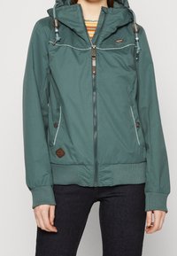 Teal windbreaker jacket with a hood, zipper closure, side pockets, and ribbed cuffs. Features a small logo patch and drawstring accents.