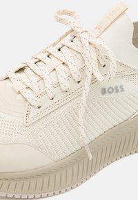 Beige kötött sportcipők texturált anyaggal, tonális részletekkel és lapos fűzőkkel. Gumi talp jellemzi, valamint a "BOSS" márkajelzés szürke színben az oldalon.