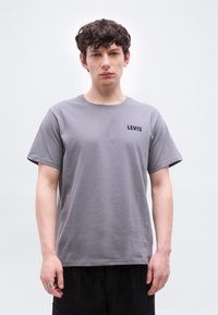 Νέος άνδρας με κατσαρά μαλλιά, που φοράει γκρι μπλουζάκι Levi's και μαύρο παντελόνι, σε απλό λευκό φόντο.