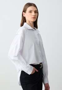 Biała koszula z guzikami o luźnym kroju, z klasycznym kołnierzykiem i długimi rękawami, zestawiona z czarnymi spodniami i czarnym paskiem.