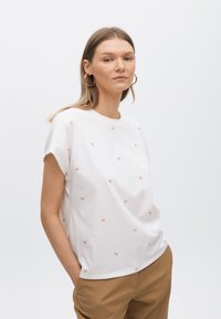 T-shirt en coton blanc avec des manches courtes retroussées, ornée de logos 'M' couleur orange clair éparpillés, associée à un pantalon beige. Design simple et décontracté.