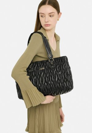 Borsa nera in tessuto con texture e motivo ondulato, caratterizzata da un design cucito, dettagli dorati e doppi manici. Il modello tiene la borsa contro uno sfondo chiaro.