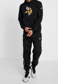 Sweat à capuche noir avec un logo viking, accompagné d'un pantalon noir avec des bandes blanches sur les côtés et des chaussures de sport blanches. Le matériau semble texturé.