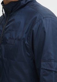 Navy blauwe nylon jas met een hoge kraag, een rits aan de voorkant en een kleine logo patch aan de linkerkant. Heeft een gladde textuur en een minimalistisch ontwerp.