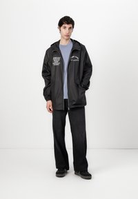 Black waterproof jacket with hood, front zipper, and printed text accents. Paired with a light blue shirt and dark denim pants.