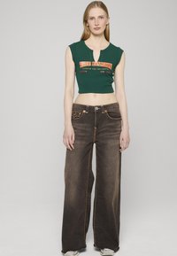 Zelené krátké tričko se oranžovým nápisem, doplněné širokými hnědými džínami se stehy v kontrastní barvě a zapínáním na knoflíky.