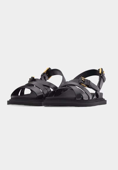 Μαύρα δερμάτινα σανδάλια με λουριά που διασταυρώνονται, αγκράφες σε χρυσό τόνο και επίπεδες σόλες, πάνω σε απλό ανοιχτό γκρι φόντο.