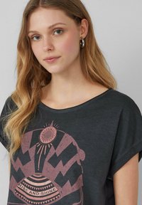 Grijze T-shirt met korte mouwen en een ronde hals. Heeft een grote, roze grafische print met geometrische patronen en de tekst "RISE AND SHINE."
