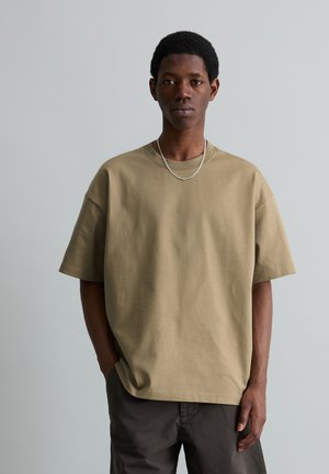 Jeune homme portant un t-shirt beige oversized, un pantalon foncé et un collier chaîne argenté, debout devant un fond gris clair uni.