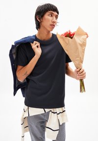 T-shirt à manches courtes bleu marine, superposé avec une veste en denim sur l'épaule, tenant un bouquet de fleurs enveloppé dans du papier kraft. Chemise rayée nouée à la taille.